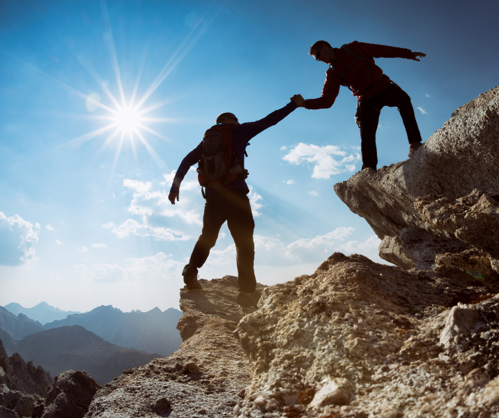 A stock image showing one person heling another reach the summit of a mountain, representing looking out for each other.