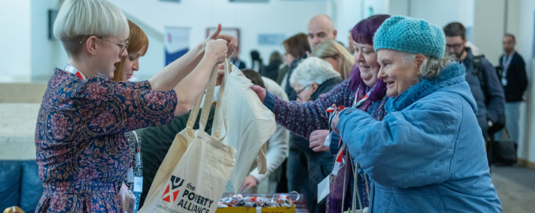 Poverty Alliance staff helping delegates register at our 2022 conference. A young woman handing over a Poverty Alliance tote bag to an older woman dressed all in blue, with a blue hat.